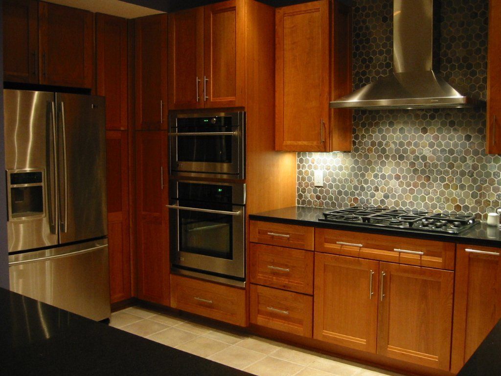 Cherry cabinetry with stacked wall ovens, a hexagon glass mosaic backsplash and black countertops
